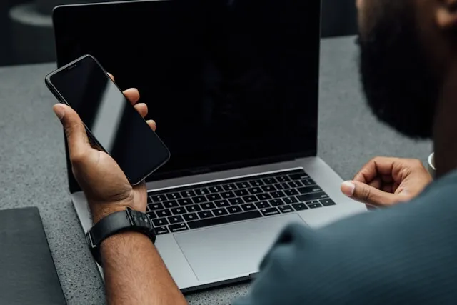 Man sitting with a laptop and holding a mobile phone, looking at the phone to make a contact or response